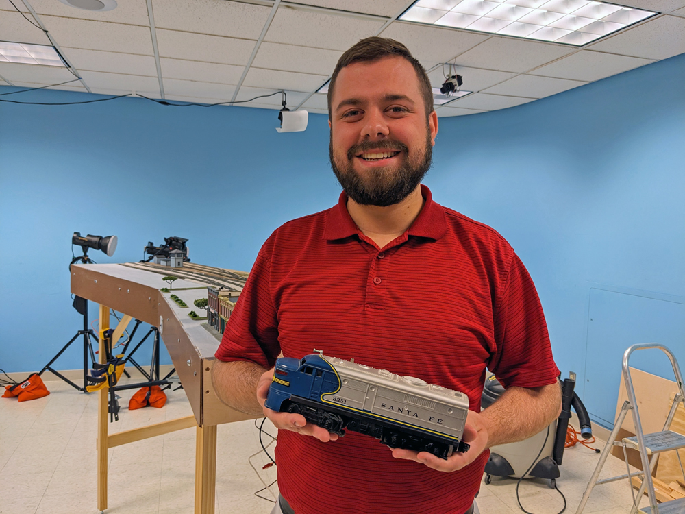 A man in a red polo shirt holds a model of a blue and silver streamlined diesel locomotive.