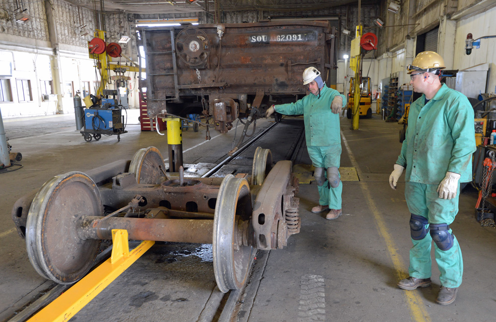 Two green-clad workers guide a rail truck out from under a gondola car