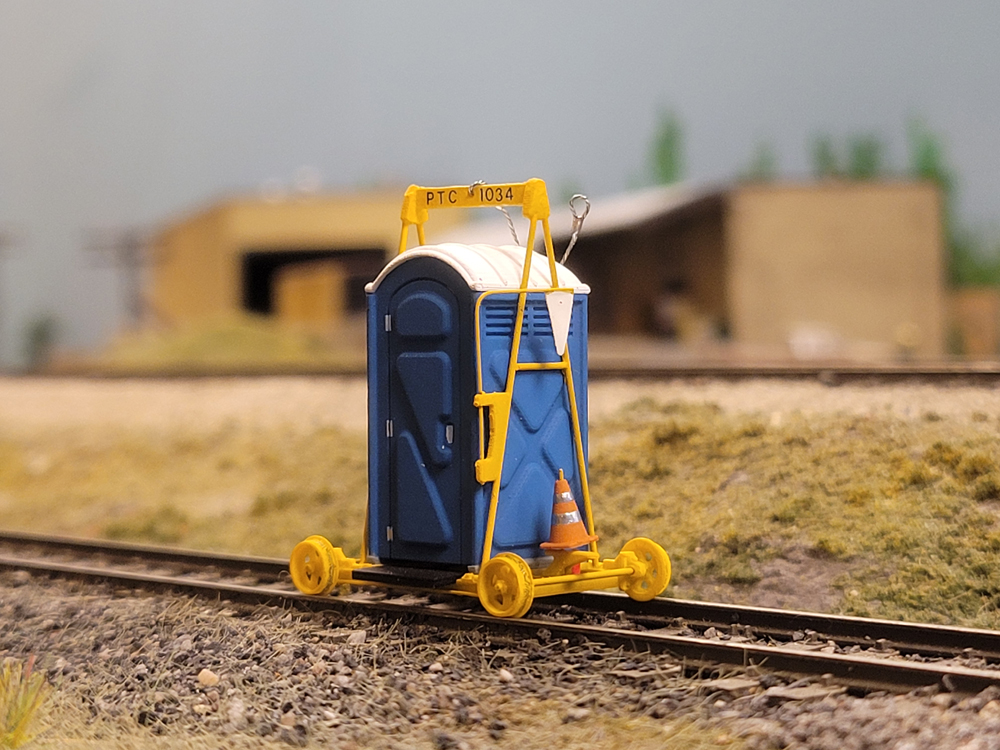 A 3-D printed model rail cart porta-potty on a model railroad layout with a rail structure in the background