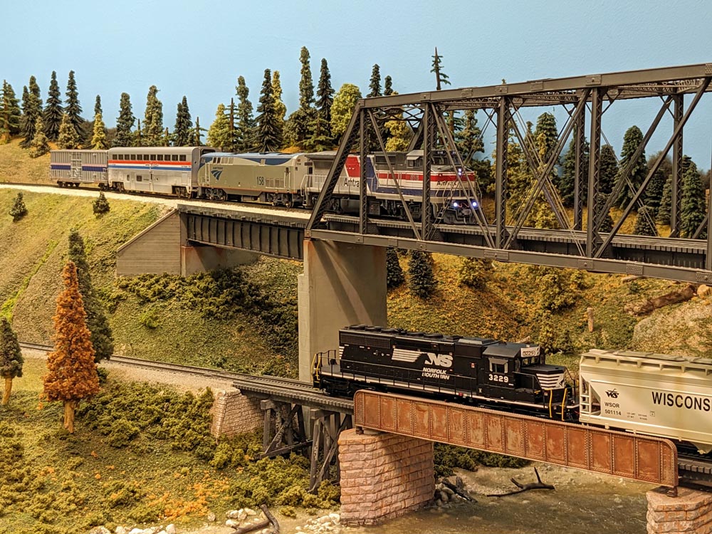 A model passenger train crosses a bridge over water and a freight train