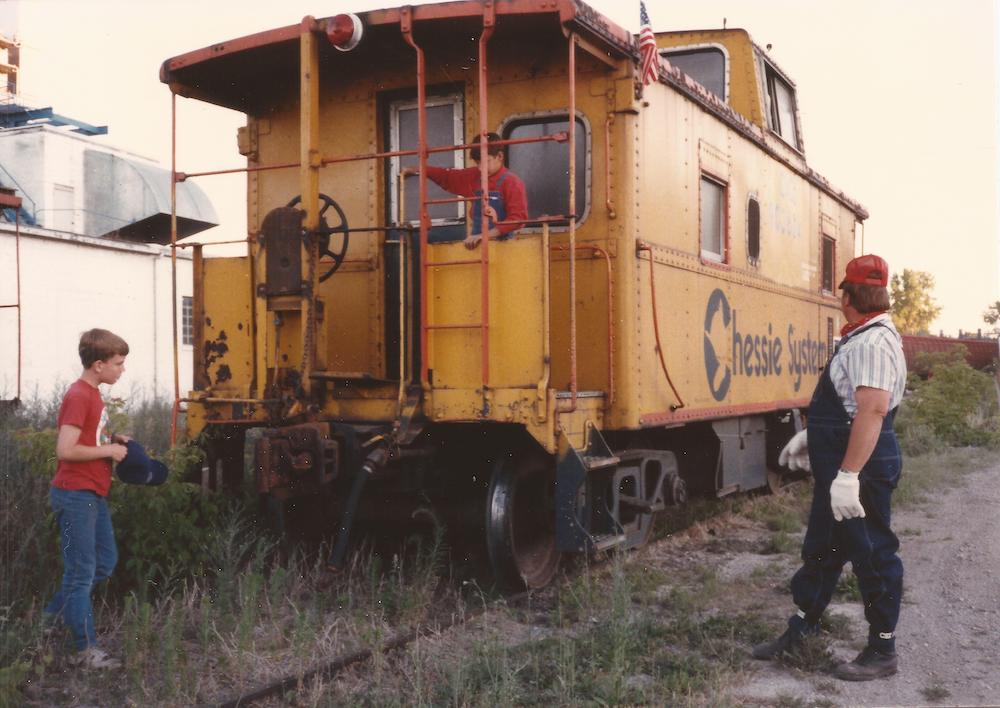 Caboose on a siding with a boy on the platform plus a second boy and conductor standing by.