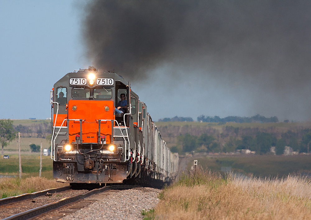 Gray locomotive with orange nose under cloud of black smoke on plains