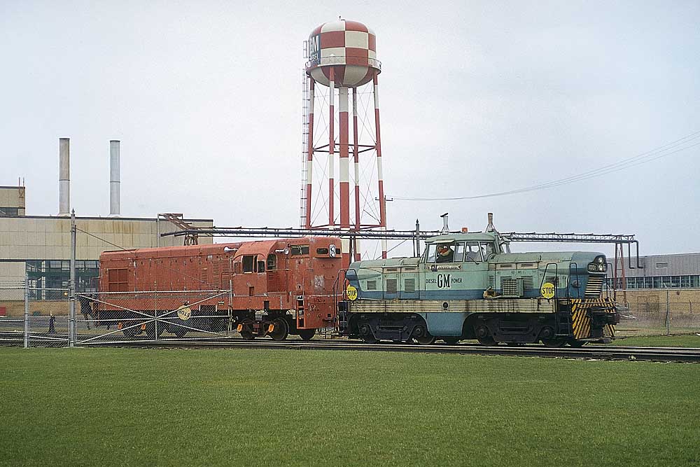 Green-and-blue centercab GMDH1 diesel-hydraulic locomotives below red checked water tower
