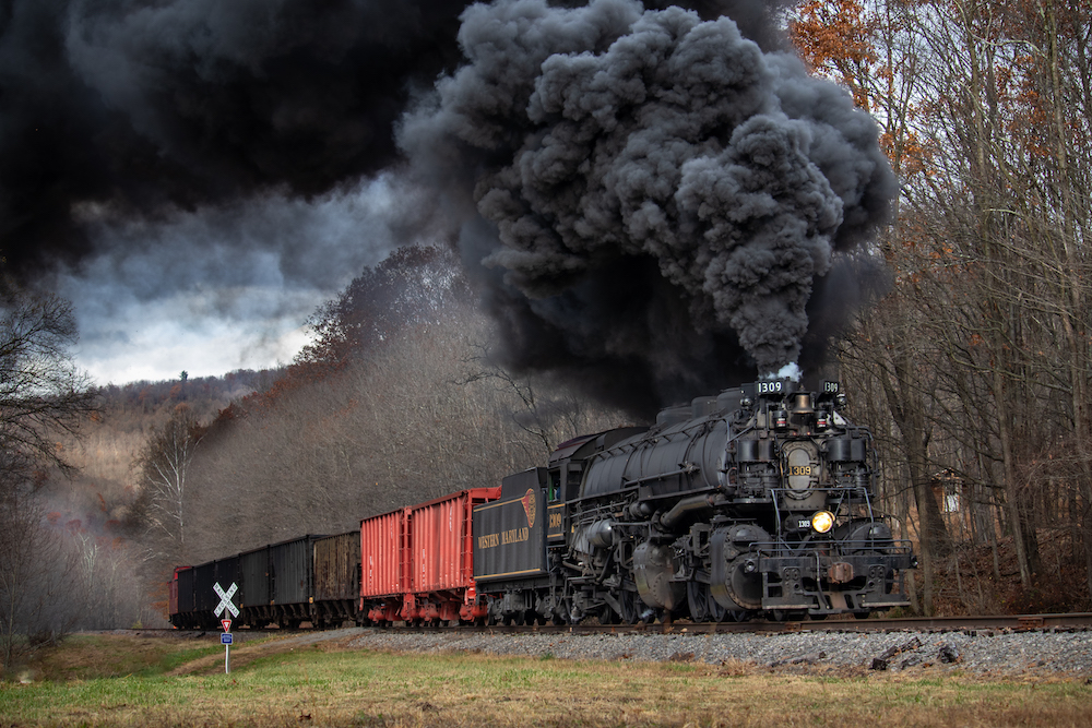 A steam locomotive rolls along under a dense black cloud of exhaust smoke