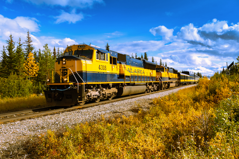 A yellow and blue locomotive rolls along under blue partly cloudy skies with golden vegetation in the foreground