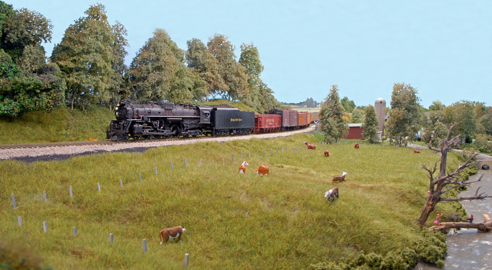 A steam locomotive pulls a freight past a field with cows on an HO scale layout