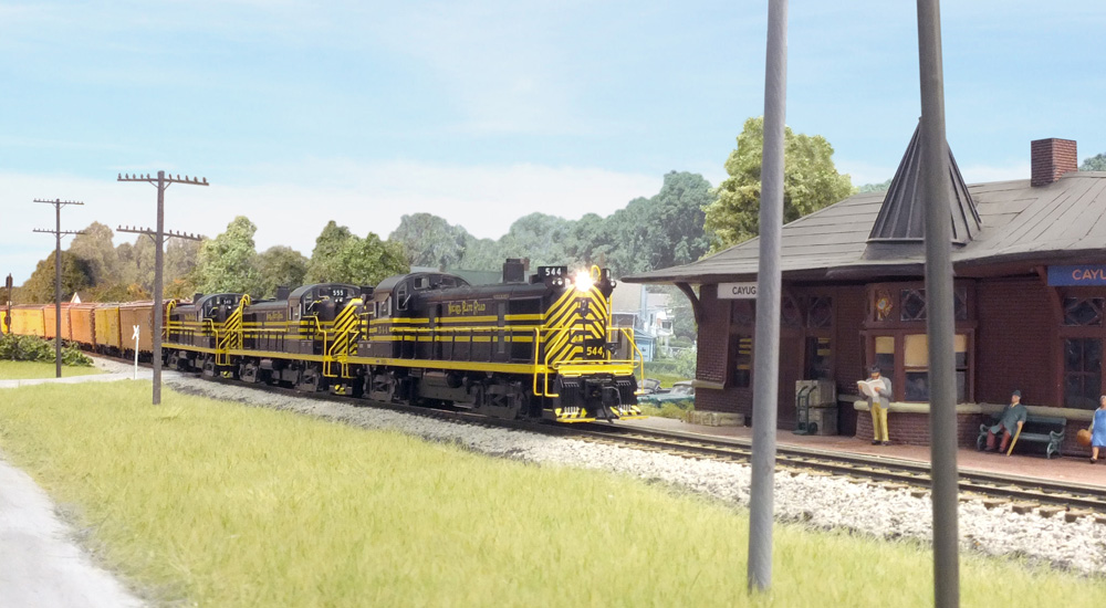 Black-and-yellow diesels lead a reefer train past a red depot