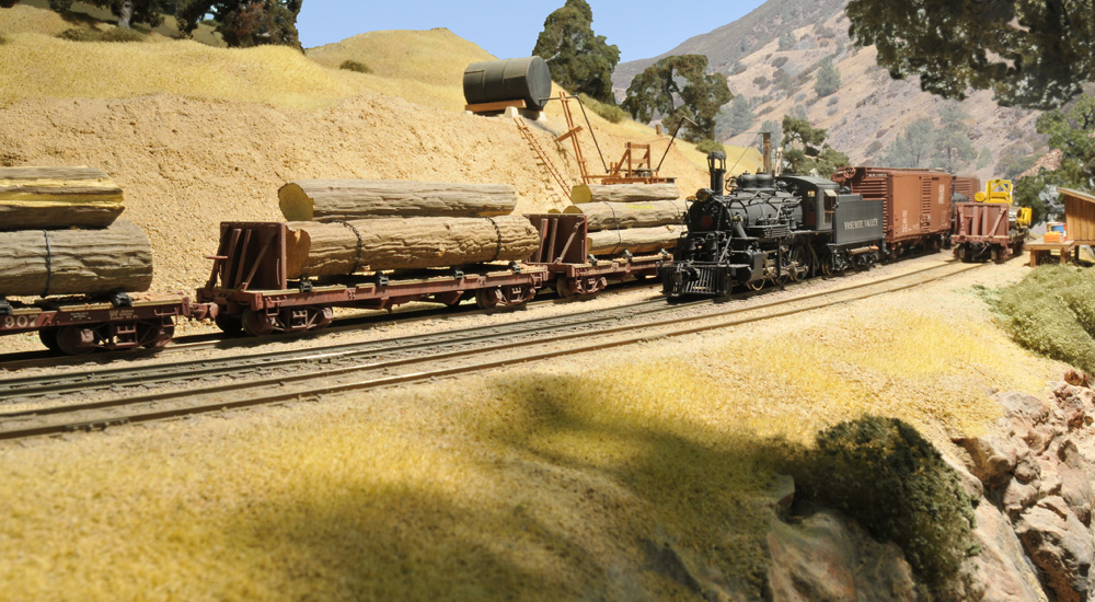 A steam locomotive takes a curve past a log train on a model railroad