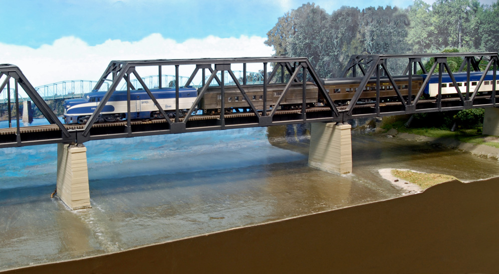 An HO scale passenger train led by a blue-and-white diesel rolls on a steel truss bridge over a river