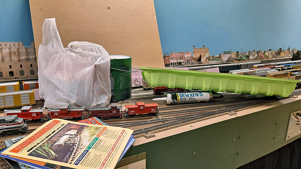 Magazines, a bag of new purchases, a sheet of cardboard, a can of paint, and a wallpaper tray mingle with freight cars in an HO scale layout’s yard.