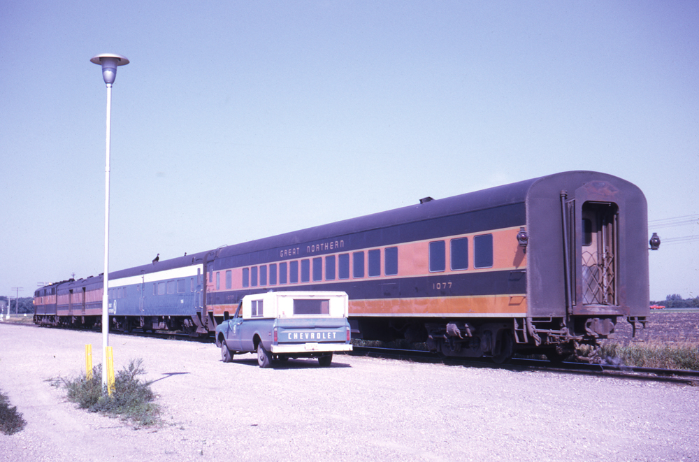 Color photo of three-car passenger train with blue truck in front of last car.