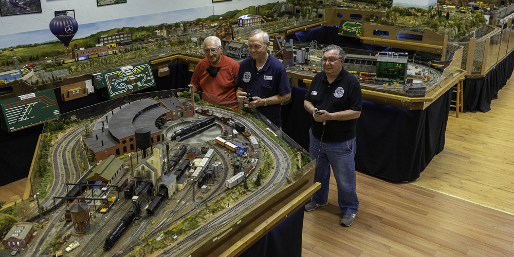 The Grand Strand Western Railroad layout: Three men standing at a model railroad layout holding controllers