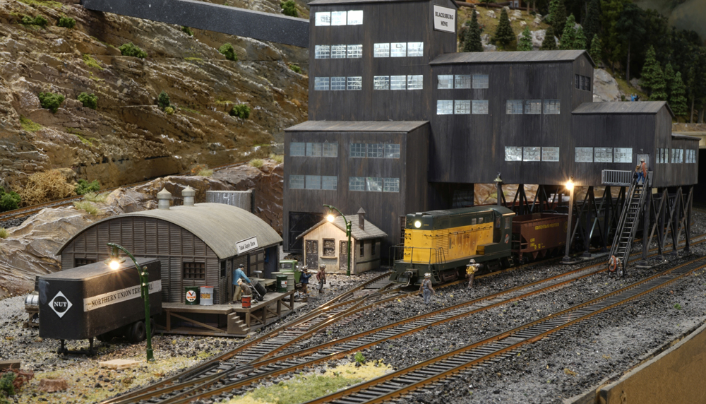 A model railroad layout with a trai passing under a large industrial structure