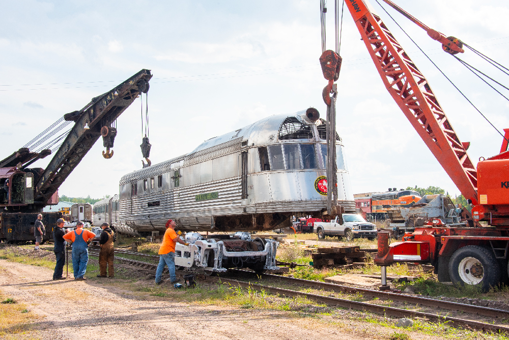 Two cranes lifting a shovel-nosed locomotive. Wisconsin Great Northern places 'Mark Twain Zephyr' back on its trucks.