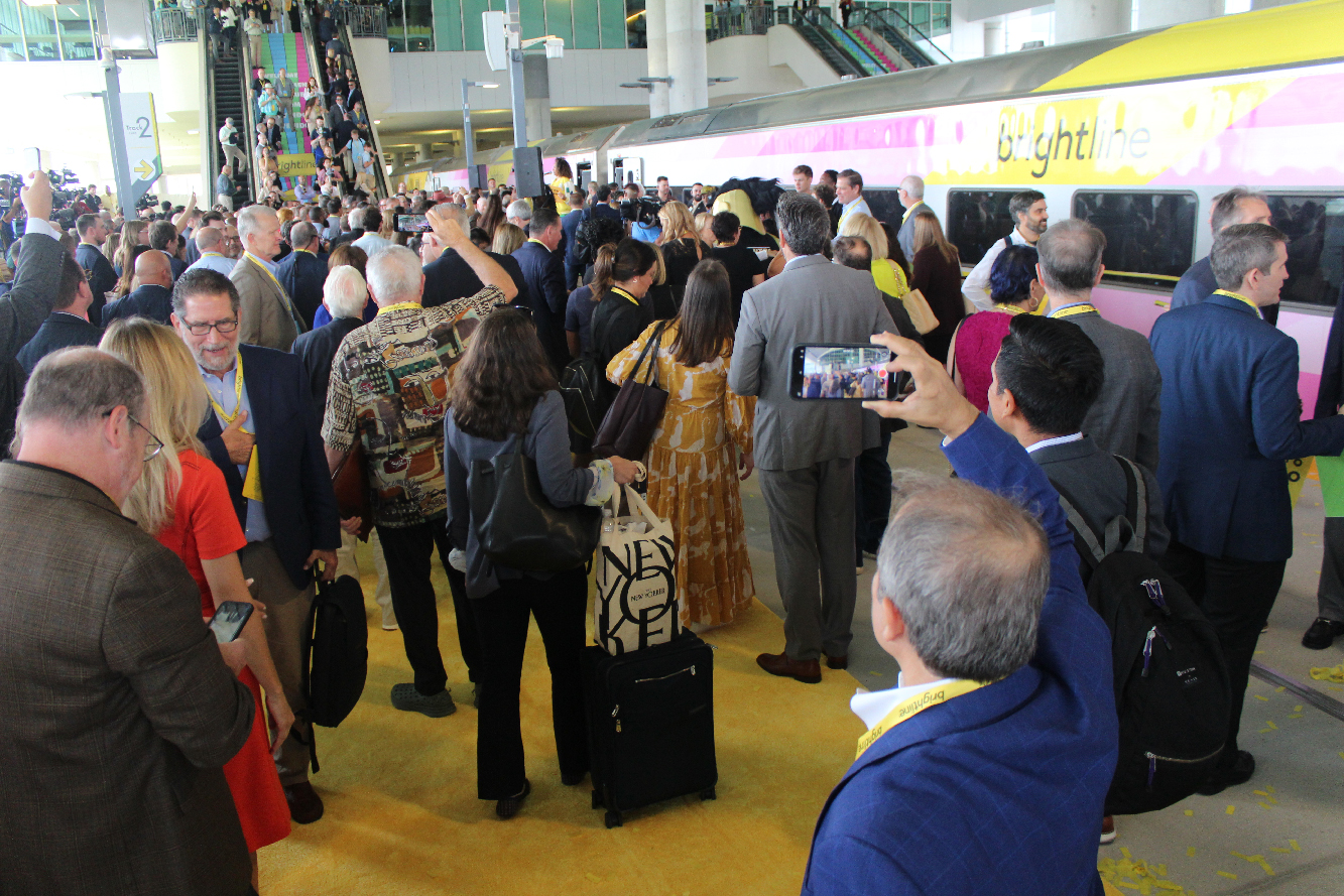 Crowd of people on station platform with pink, yellow, and white passenger train.