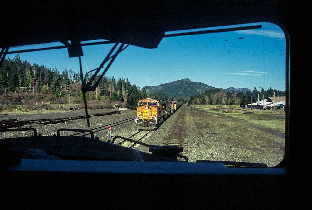 An orange BNSF engine approaches through tree on either side of the tracks with a mountain in the background.