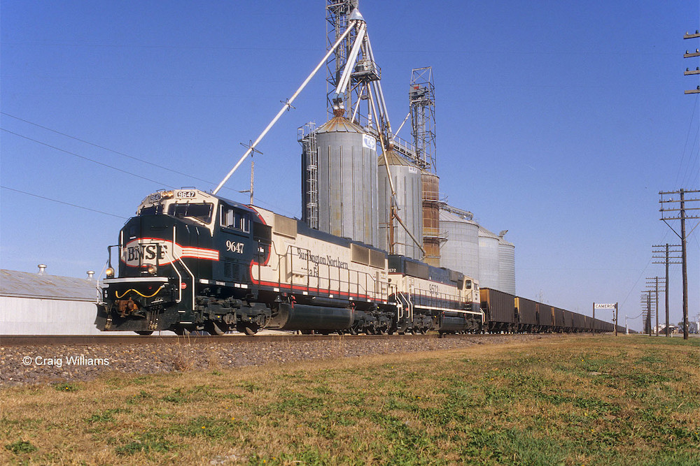 Cream-colored diesel locomotives pass a green field in the foreground and the tall silver bins of a grain elevator in the background