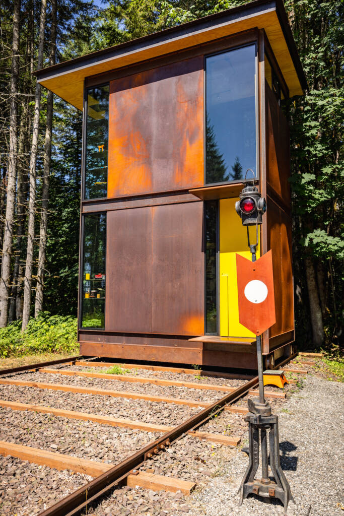 A two-story office structure made of glass and unpainted steel sheets sits on a wide-gauge track