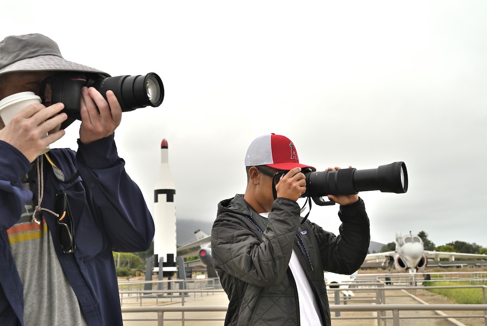 Two people, holding cameras and wearing hats under a gray sky