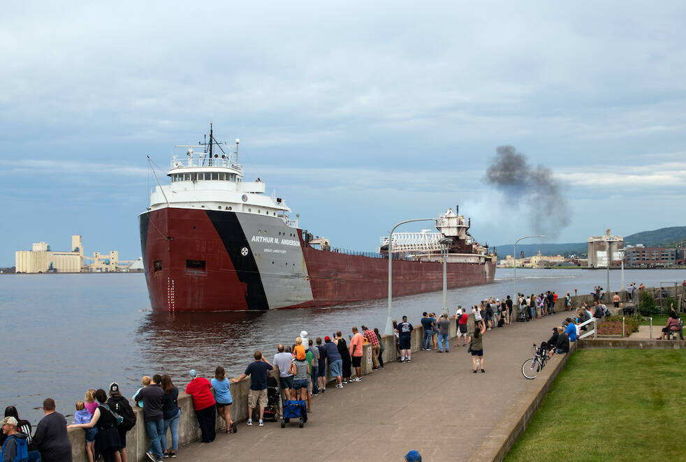 A crowd of onlookers line the railing of a pier while a red and white ship eases past on a cloudy day