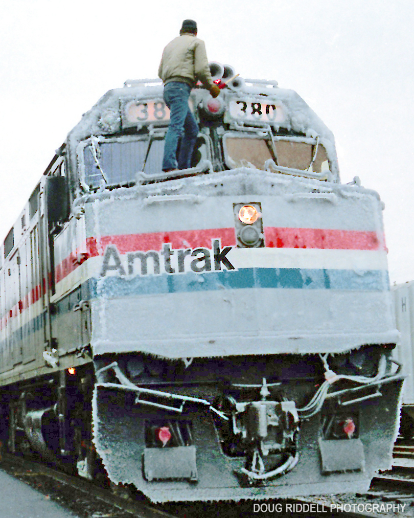 A crewman standing on the hood of a silver locomotive chips accumulated ice off the engine under gray skies