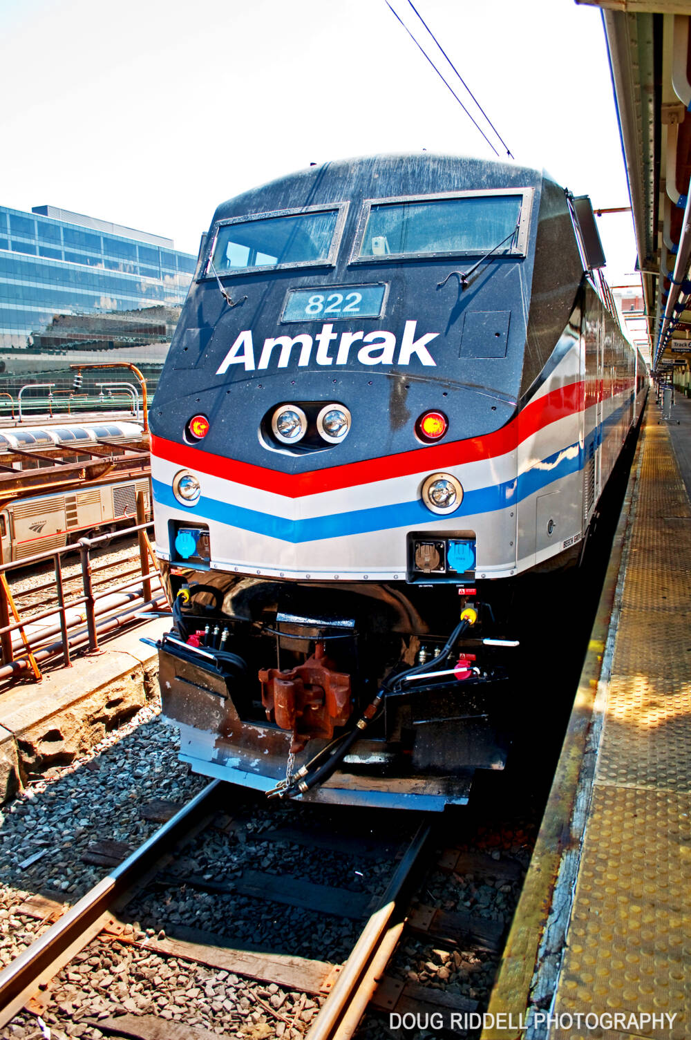 A modern passenger diesel painted silver, with a black nose and red, white and blue stripes at a passenger station platform
