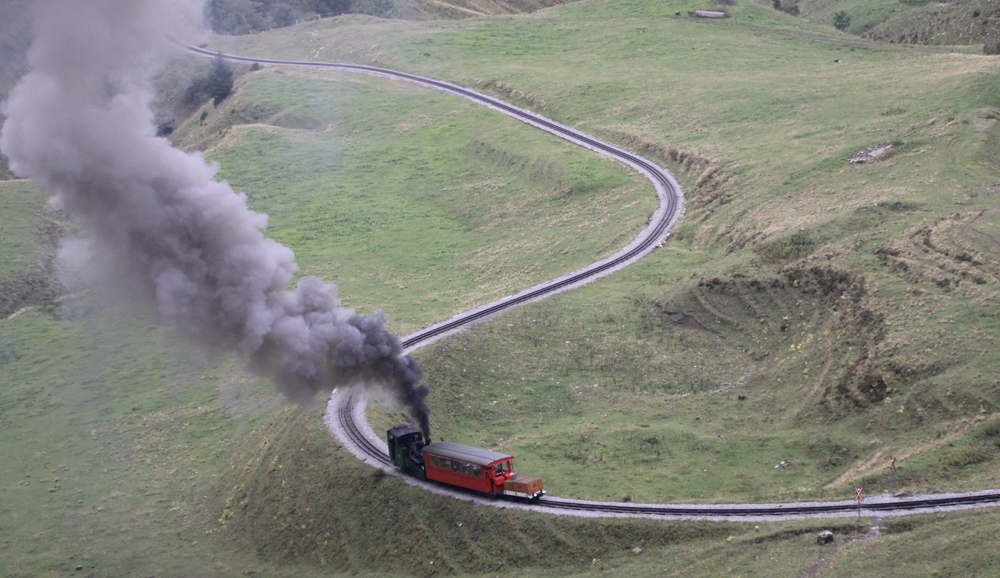 Steam locomotive and one car on curving line on mountainside, as seen from above