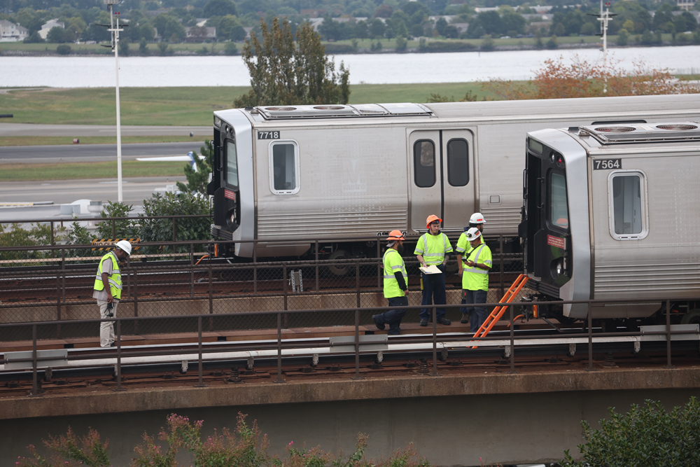 Men in high-visibiilty clothing stand near derailed rapid-transit equipment as another train passes