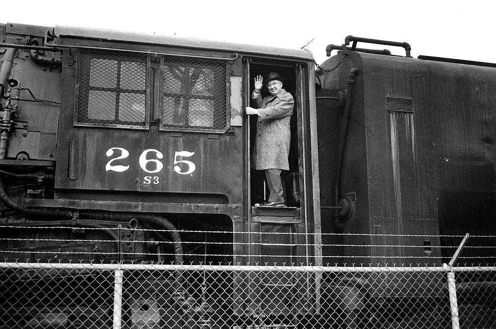 a man in an overcoat waves from the door of a steam locomotive in a black and white image