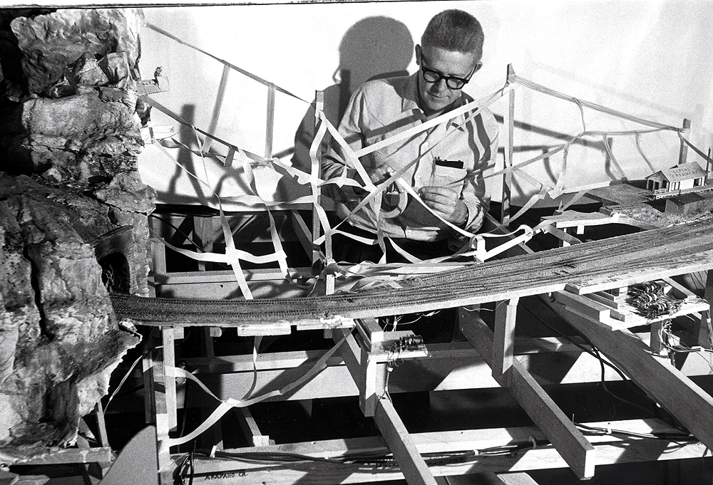 A man strings masking tape between supports while standing in the midst of model railroad benchwork in this black and white image