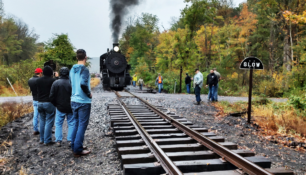 Small group of people watching a black steam locomotive approach on the tracks.