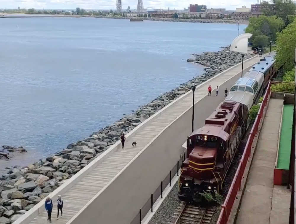 Maroon diesel locomotive pulling passenger train along a lake.