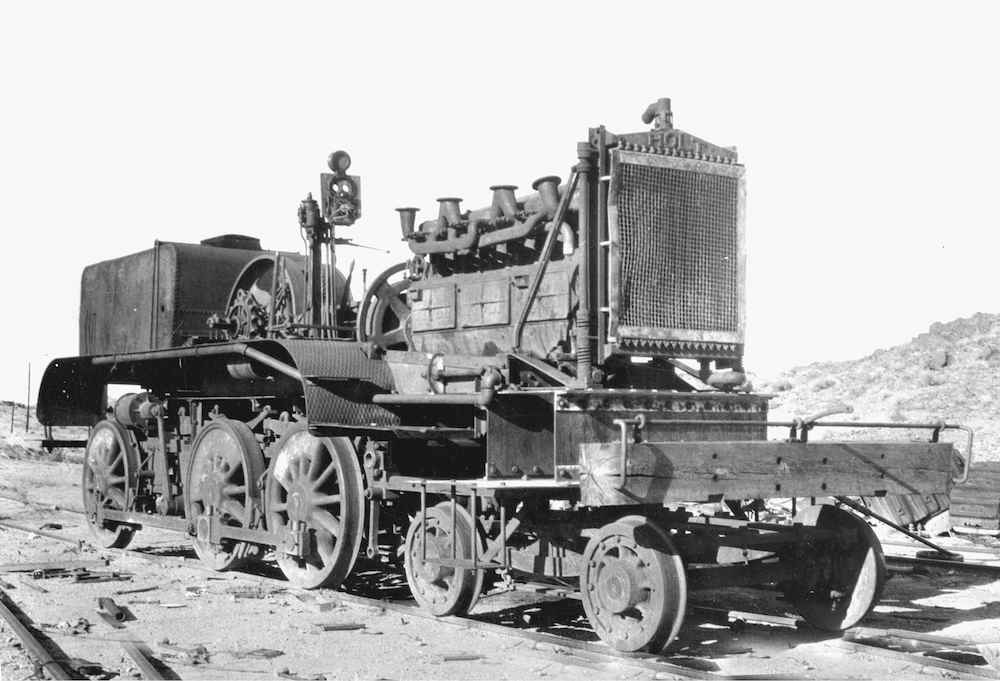A vintage black and white photo shows a tractor engine atop a former steam locomotive frame and wheels