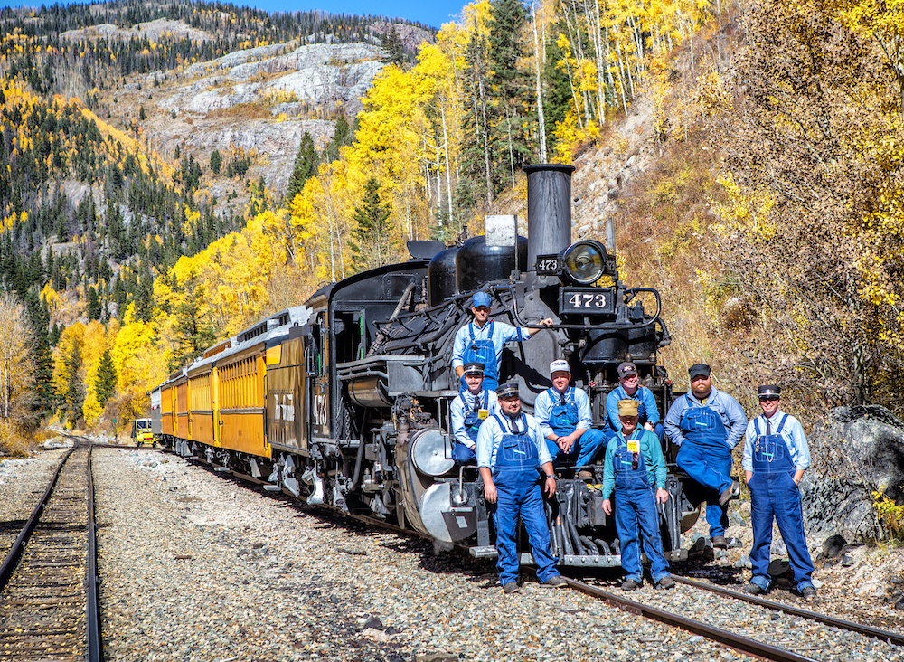 Dressed in blue overalls, a railroad crew poses in front of a black steam locomotive pulling yellow passenger cars
