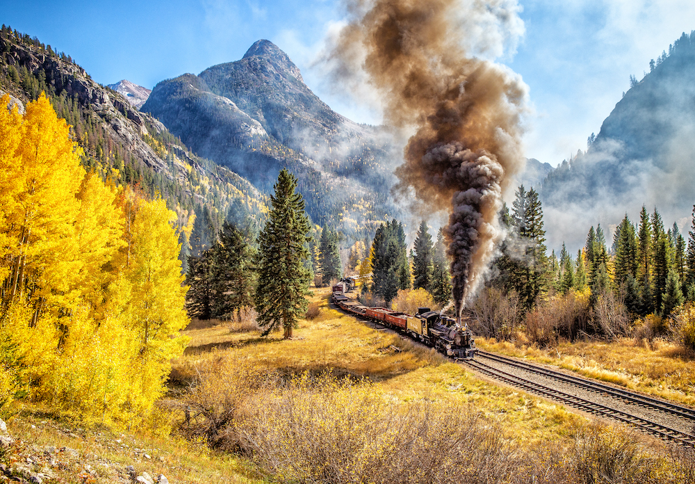 Tall rocky peaks surround a steam train making its way through a tree-filled mountain valley