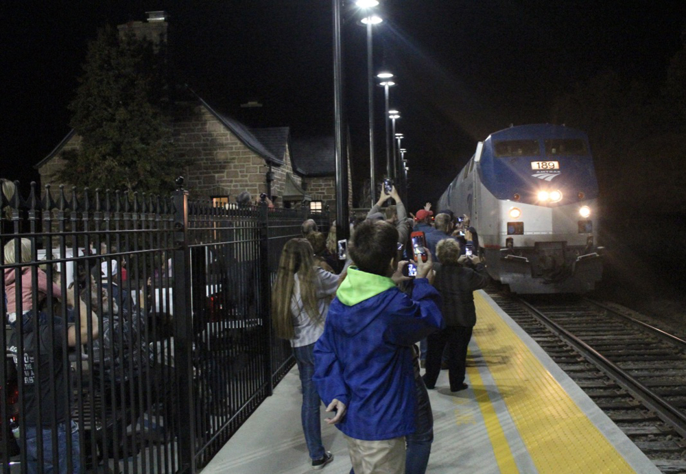 People, many with cellphones taking pictures, crowd station platform as passenger train arrives at night