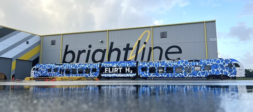 Blue and white hydrogen trainset with elaborate pattern on body next to building with large Brightline sign