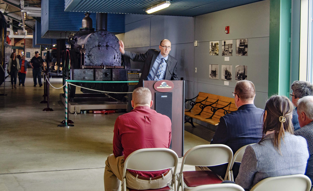 Man speaking at podium gestures toward antique locomotive in background.