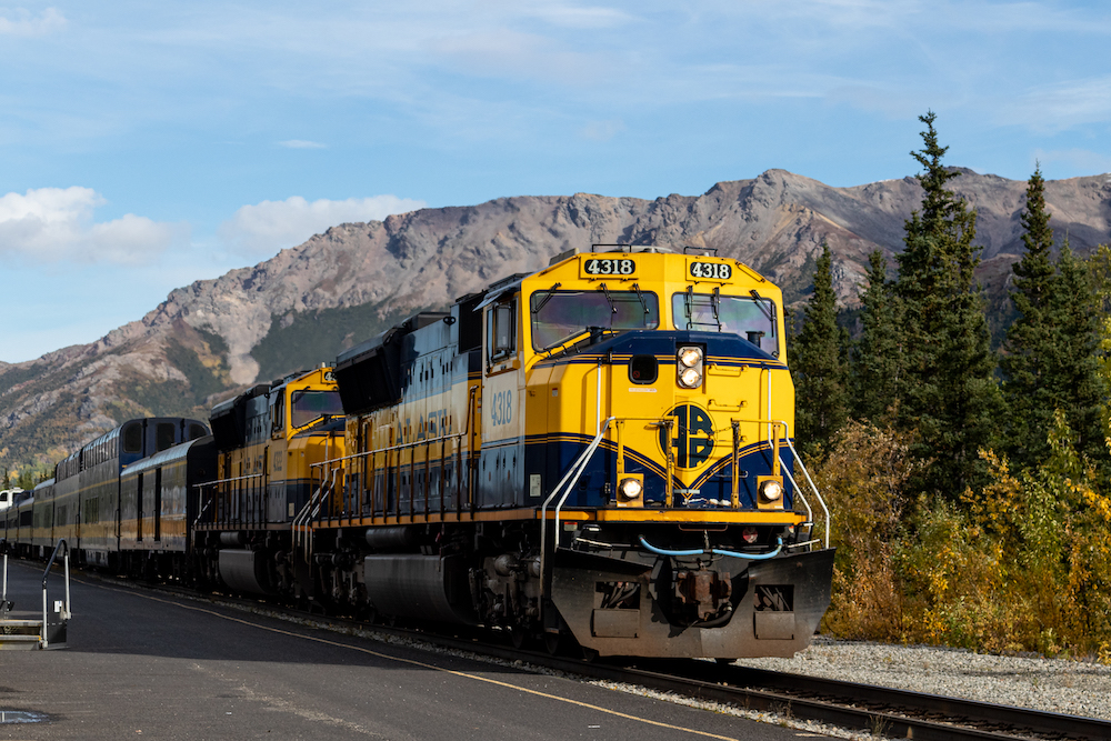 With gray mountain peaks above it, a yellow and blue passenger train pulls in to a station