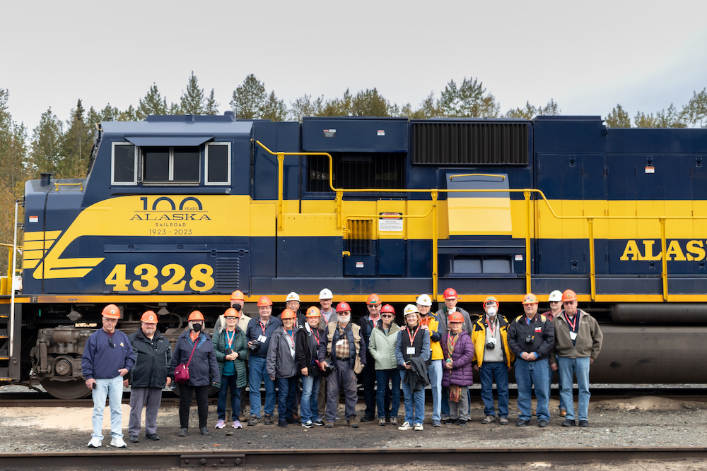 Two dozen people in hardhats stand alongside a blue and yellow diesel locomotive