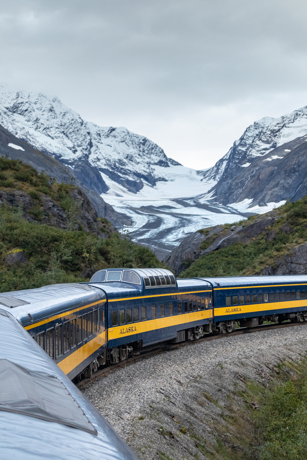 Passenger train cars curve past a backdrop of snow-covered mountains.