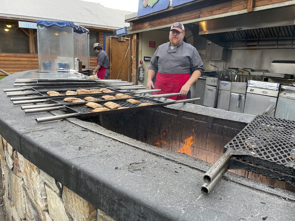 A cook in a red apron watches over racks of salmon filets on an open fire.