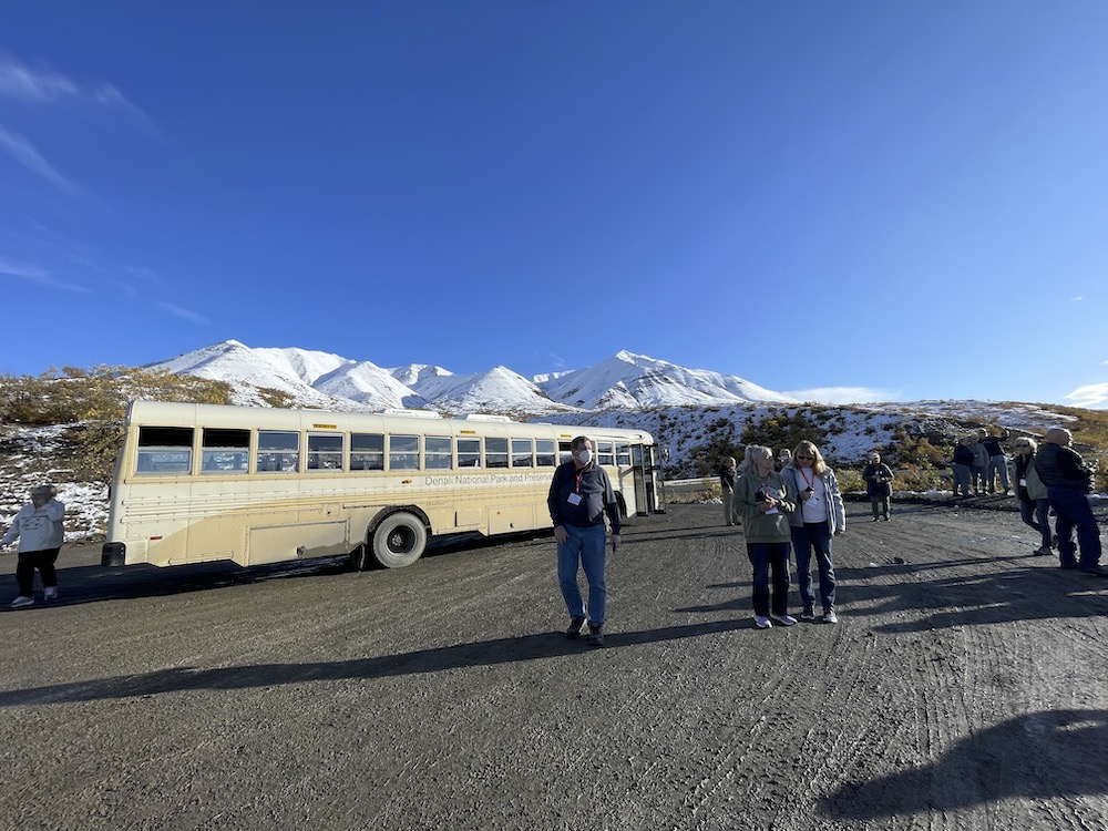 A beige school bus is parked on a gravel road while its passengers mill around. A snow-coereed mountain range is the background.
