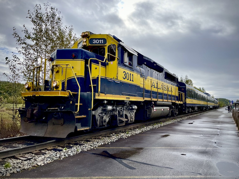 A blue with yellow stripe diesel locomotive and short train next to a rain-soaked platform