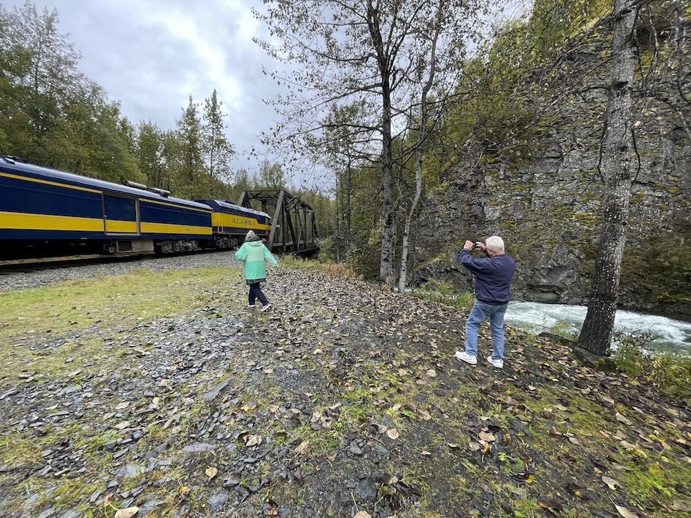 Two people take photos of a stopped passenger train next to a rock cliff face and whitewater stream