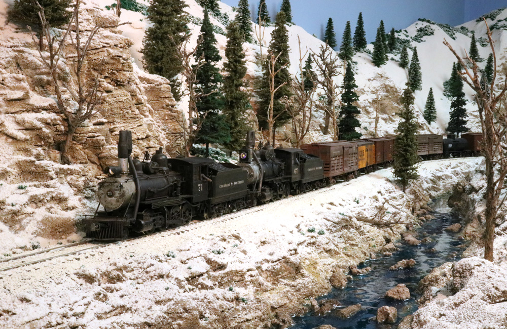 Two steam engines pull a freight alongside a creek and through a rock cut