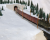 A green boxcab electric locomotive pulls a string of boxcars from a tunnel and through a snowy landscape