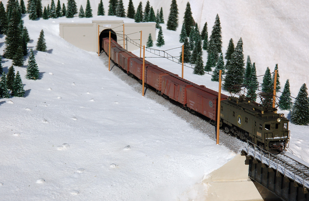 A green boxcab electric locomotive pulls a string of boxcars from a tunnel and through a snowy landscape