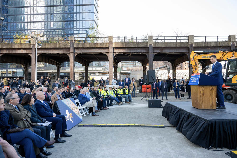 Man speaking at podium in front of large crowd outdoors, with overpass in background