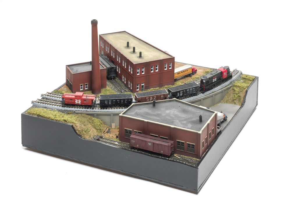 Small model train scene with three red-brick buildings, tall brick chimney, two gray railroad tracks, and a short train of black and brown coal hopper cars.